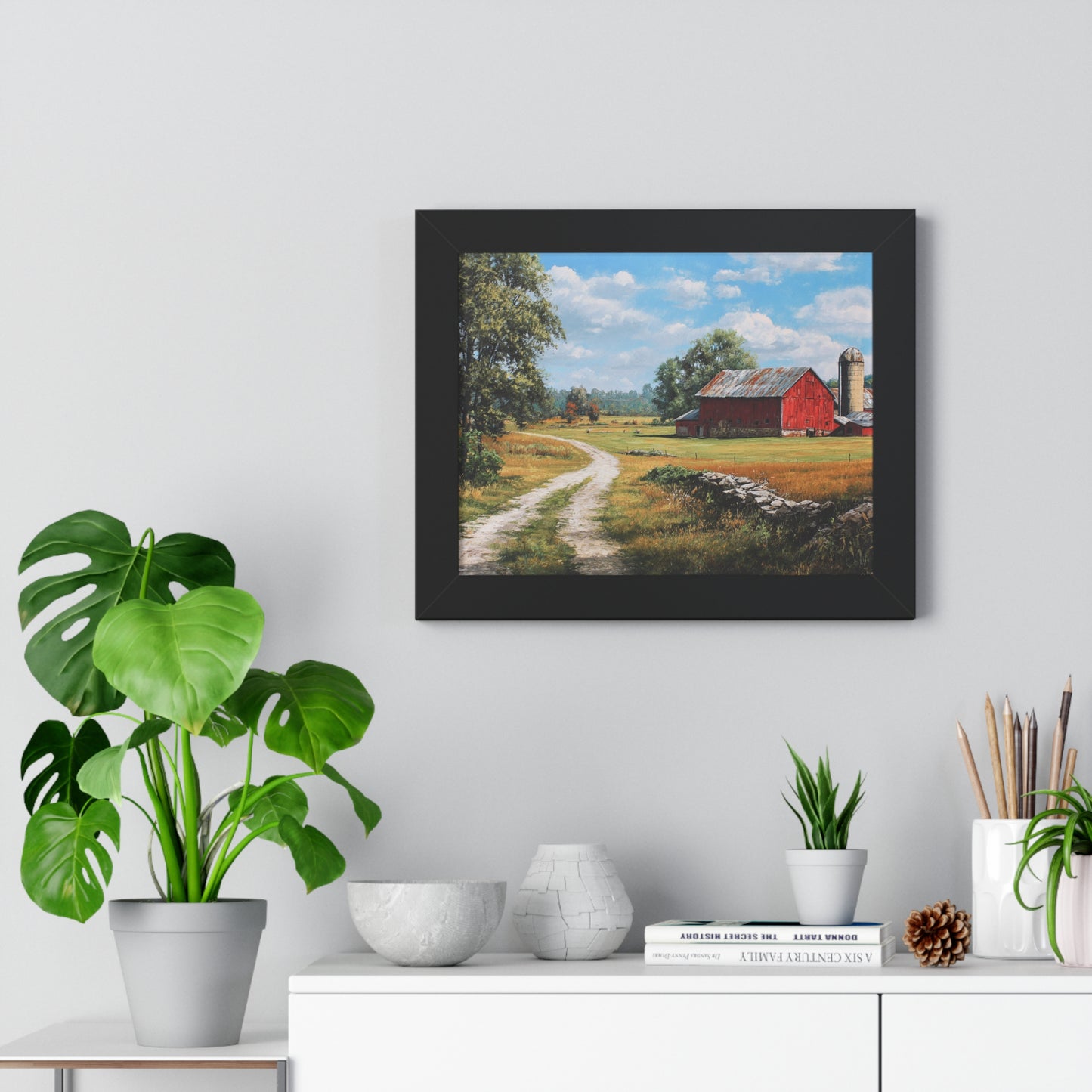 Framed painting of a rural landscape with a red barn on a wall above a white cabinet with decorative items.